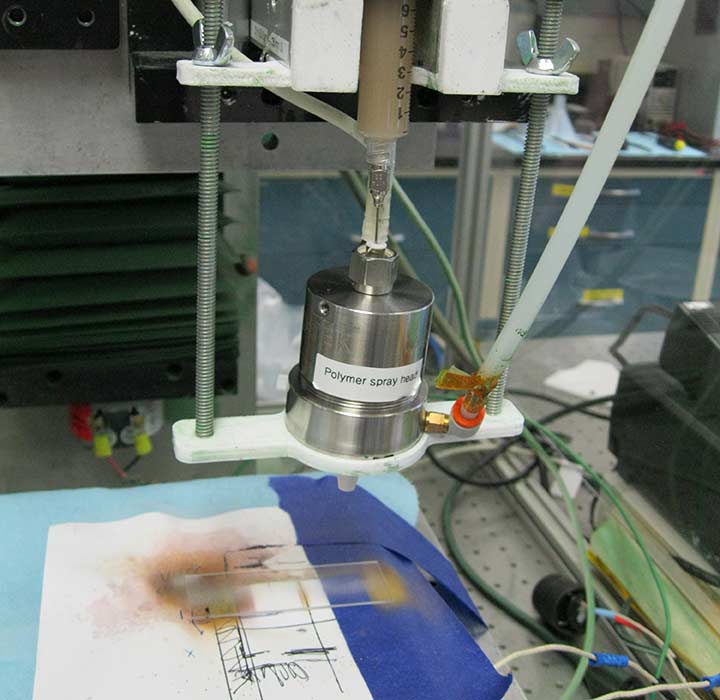 A polymer spray head in a lab spraying a rectangular piece of glass